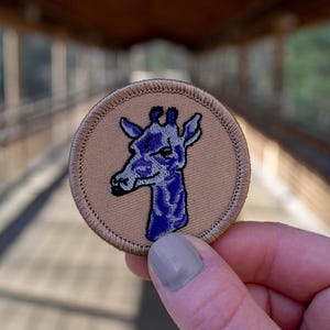 May include: A brown embroidered patch with a purple giraffe head design. The giraffe has a light blue eye and a light blue and purple patterned body.