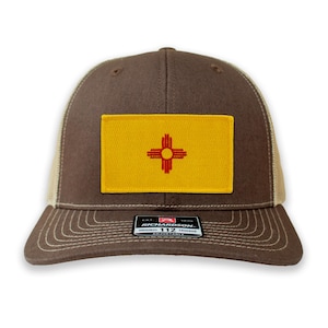 May include: Brown and beige trucker hat with a yellow and red New Mexico state flag patch. The hat has an adjustable strap with the text "Richardson 112 Original Trucker Adjustable" on it.