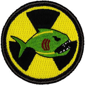 May include: A yellow and black radiation symbol with a green fish with red gills and white teeth inside. The fish is facing right and has a menacing expression.