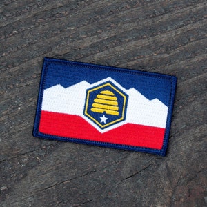 May include: A blue, white, and red embroidered patch with a yellow beehive and a white star in the center. The patch is on a dark wood surface.