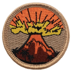 May include: A circular embroidered patch featuring a volcanic eruption with a dark brown mountain base, bright orange lava, and a yellow explosion against a tan background. The design is framed by a thick, tan stitched border.