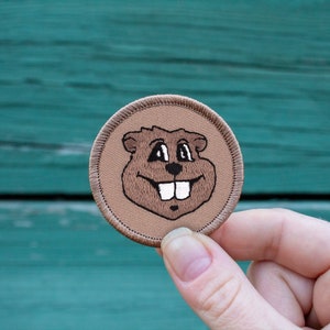 May include: A brown embroidered patch with a cartoon image of a smiling groundhog with white teeth. The patch is circular and has a brown stitched border.