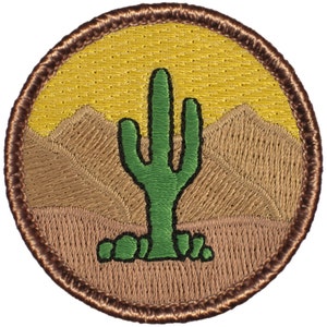 May include: A brown embroidered patch with a green cactus in the center, surrounded by brown mountains and a yellow sky.