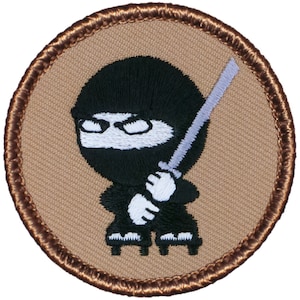 May include: Embroidered patch featuring a cartoon ninja in a black mask and outfit, holding a silver sword. The ninja is set against a tan background with a brown stitched border. The design is a playful and stylized representation of a ninja.