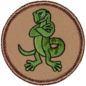 May include: A green embroidered patch with a cartoon lizard with its arms crossed. The lizard is standing on a tan background with a brown border.