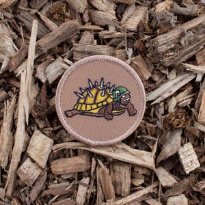 May include: A brown embroidered patch with a cartoon turtle wearing a green helmet and yellow spiked shell.