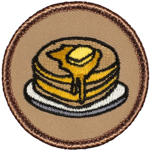 May include: A brown embroidered patch with a yellow and brown pancake with a yellow pat of butter on top. The pancake is on a white plate with a gray rim.