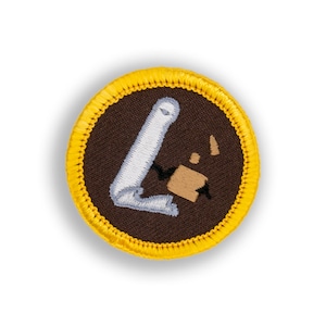 May include: A brown and yellow embroidered scout patch with a white and gray image of a rolled up scroll with a small brown box on top.