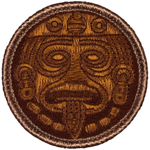 May include: A brown embroidered patch with a stylized tiki mask design. The mask has a large nose, wide eyes, and a wide mouth.
