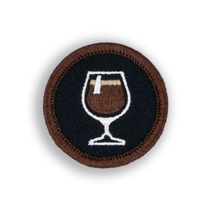 May include: A brown embroidered patch with a white outline of a glass of coffee. The patch is circular and has a brown border.