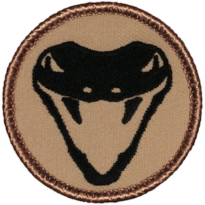 May include: A round, tan fabric patch with a brown embroidered border. The patch features a black embroidered image of a cobra's head with fangs.