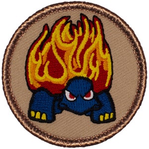 May include: A round embroidered patch with a brown border. The patch features a cartoon turtle with flames on its back. The turtle has red eyes and a blue shell. The flames are red, yellow, and orange.