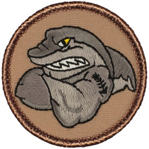 May include: A brown embroidered patch with a gray cartoon shark with a yellow eye and a white mouth. The shark is flexing its bicep.