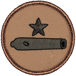 May include: A brown embroidered patch with a black star and a gray cannon. The patch is surrounded by a brown border.
