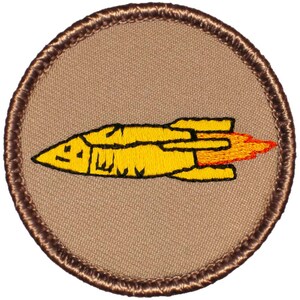 May include: A brown embroidered patch with a yellow rocket ship with orange flames. The rocket ship is flying to the right.