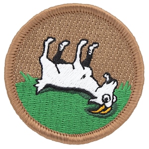 May include: Embroidered patch featuring a cartoon goat lying upside down on green grass. The goat is white with black hooves and a yellow beard. The patch has a brown border and a tan background.