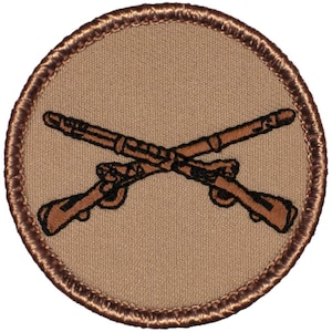 May include: A brown embroidered patch with a circular shape and a brown border. The patch features two crossed rifles in the center.
