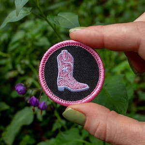May include: A round, embroidered patch featuring a pink cowboy boot on a black background, encircled by a pink border. The patch is held against a backdrop of green foliage and purple flowers.