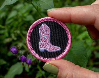 May include: A round, embroidered patch featuring a pink cowboy boot on a black background, encircled by a pink border. The patch is held against a backdrop of green foliage and purple flowers.