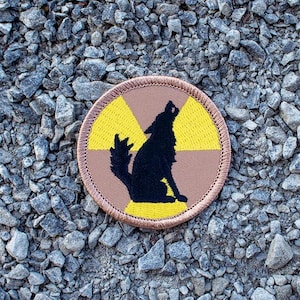 May include: A brown and yellow embroidered patch with a black silhouette of a wolf howling at the moon. The patch has a yellow radiation symbol in the background.