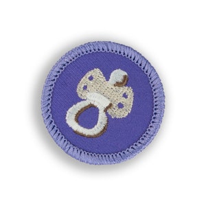 May include: A purple embroidered patch with a white pacifier design. The pacifier has brown details.