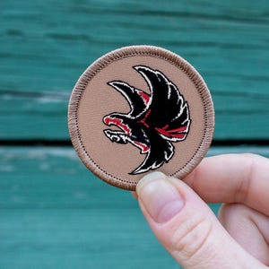May include: A round, tan embroidered patch with a black and red eagle design. The eagle is in flight with its wings spread wide.