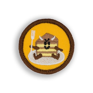 May include: A brown and yellow embroidered patch with a cartoon cake character holding a fork. The cake character has a smiling face and horns. The patch is surrounded by a brown border.