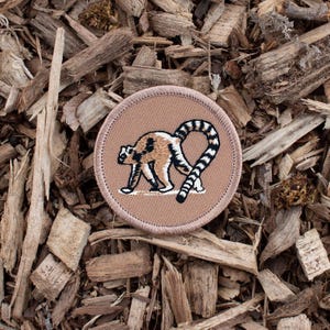 May include: A circular patch featuring a lemur design. The lemur is depicted in shades of brown, tan, and white, with a distinctive striped tail. The patch has a tan background and a stitched border, set against a backdrop of wood chips.