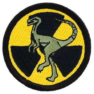 May include: A yellow and black embroidered patch with a green dinosaur inside a radiation symbol.