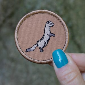 May include: A brown embroidered patch with a white and gray weasel standing on its hind legs.