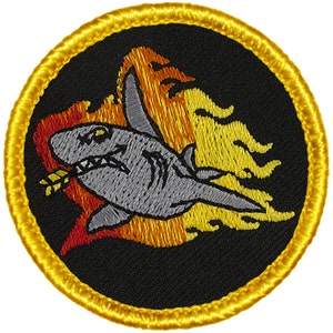 May include: A round embroidered patch with a yellow border. The patch features a gray shark with a red and orange flame behind it. The shark is biting a yellow arrow.