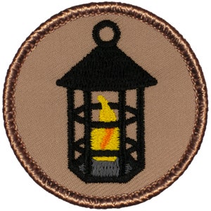 May include: A brown and black embroidered patch with a lantern design. The lantern is black with a yellow flame inside. The patch has a brown border.