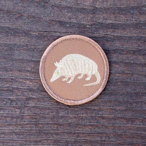 May include: A brown embroidered patch with a white armadillo on a brown background.