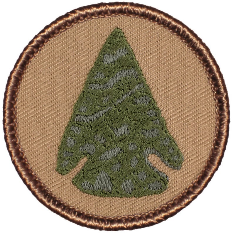 May include: A brown and tan embroidered patch with a green arrowhead design. The arrowhead is a camouflage pattern.