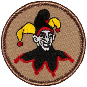 May include: A brown embroidered patch with a jester wearing a black and red hat with yellow and red bells. The jester has a white face with a black beard and a mischievous smile.