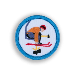 May include: A blue and white embroidered patch featuring a skier in a red hat, orange jacket, and blue pants, skiing down a slope with a ski pole in their hand. The patch has a yellow and black design in the bottom center.