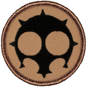 May include: A brown and black embroidered patch with a stylized skull design. The skull has two large eye sockets and a pointed crown.