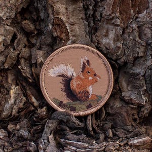 May include: A brown embroidered patch with a squirrel on a tan background. The squirrel is facing right and has a bushy tail.