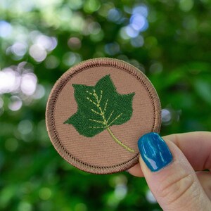 May include: A round, embroidered patch featuring a green leaf design on a tan background. The leaf has detailed stitching, and the patch has a brown border. The patch is held in a hand with blue nail polish.