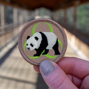 May include: A brown embroidered patch with a green biohazard symbol and a black and white panda bear inside.