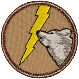 May include: A brown embroidered patch with a yellow lightning bolt and a gray bear's head. The bear is facing the lightning bolt and appears to be sniffing it.