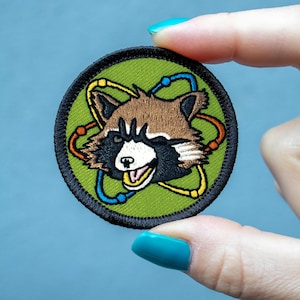 May include: Embroidered patch featuring a cartoon raccoon with a smiling expression, set against a green background. The raccoon is surrounded by a yellow, orange, and blue atom design, with a black border. The patch is round.