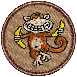 May include: A brown embroidered patch with a cartoon monkey holding two yellow bananas. The monkey is smiling with a large toothy grin. The patch is circular and has a brown border.