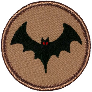 May include: A brown embroidered patch with a black bat with red eyes. The bat is flying with its wings spread out.