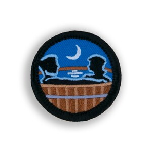May include: A round, embroidered patch with a blue background depicting two figures sitting in a wooden tub under a crescent moon. The tub has brown wooden slats and a light blue interior. The figures are black silhouettes.