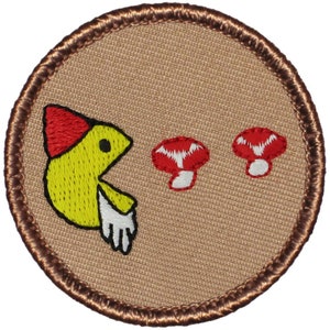 May include: A brown and white embroidered patch with a yellow Pac-Man character wearing a red hat and eating two red mushrooms.
