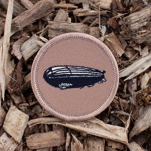 May include: A brown embroidered patch with a black and white airship design. The patch is circular and has a white stitched border.