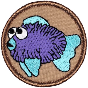 May include: A purple and blue embroidered fish patch with large eyes. The patch is circular and has a brown border.
