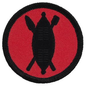 May include: A circular embroidered patch featuring a vibrant red background with a black border. In the center, a black silhouette depicts a traditional shield positioned vertically, crossed by a spear and a club, creating a bold graphic design.