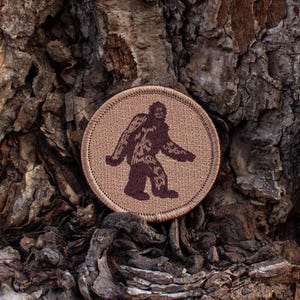 May include: A brown embroidered patch with a brown outline, featuring a brown silhouette of Bigfoot walking on a tan background.